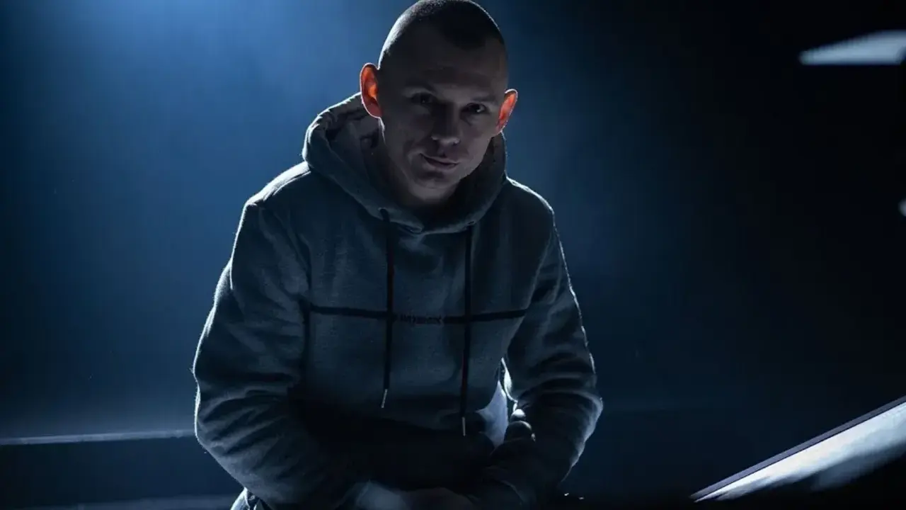 Młody mężczyzna w szarej bluzie z kapturem, z ogoloną głową, wygląda jak jeden z popularnych raperów, gotowy do występu.