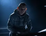 Młody mężczyzna w szarej bluzie z kapturem, z ogoloną głową, w mrocznym świetle. Jego spojrzenie sugeruje opowieść o polskim rapie.