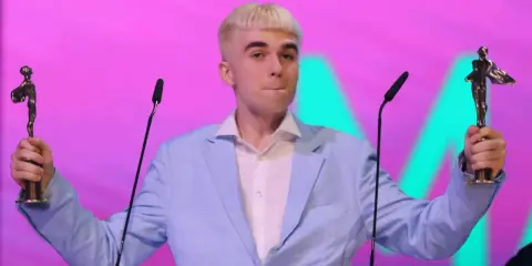 Mc Mata z dwoma statuetkami na scenie, w jasnoniebieskiej marynarce i białej koszuli.