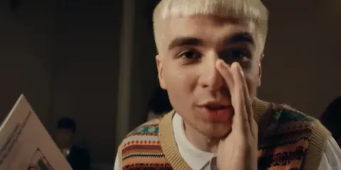 Młody mężczyzna z blond fryzurą, w kamizelce z kolorowym wzorem, trzyma rękę przy ustach, jakby coś szeptał. Obok niego leży blok mata tekst.