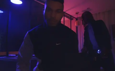 Dwóch mężczyzn w ciemnym pomieszczeniu, oświetlonym niebieskim i różowym światłem. Jeden z nich, 730 huncho, ma na sobie bluzę z logo Nike.