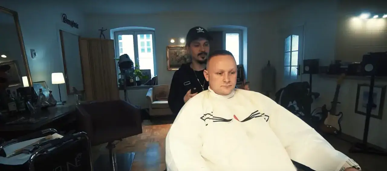 Fryzjer strzyże klienta w salonie. W tle widać gitary i lustro. Wiek Bajorsona nie jest widoczny, ale jego fryzura jest krótka.